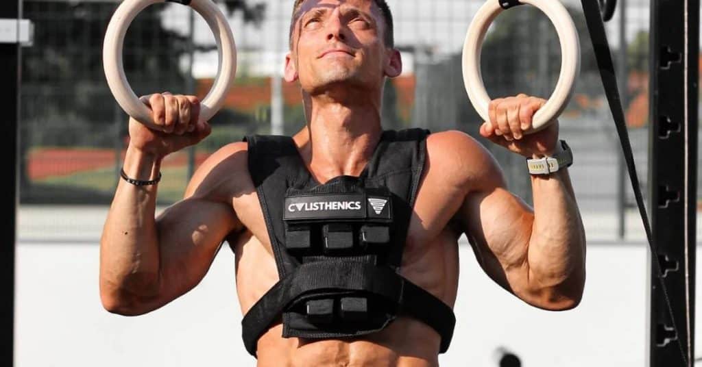 Muscular men is doing ring pull-ups with a weighted vest