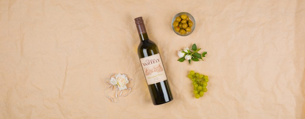 Fles Sauvignon Blanc liggend met schaalt olijven, een druiventros, bloesem op een beige ondergrond
